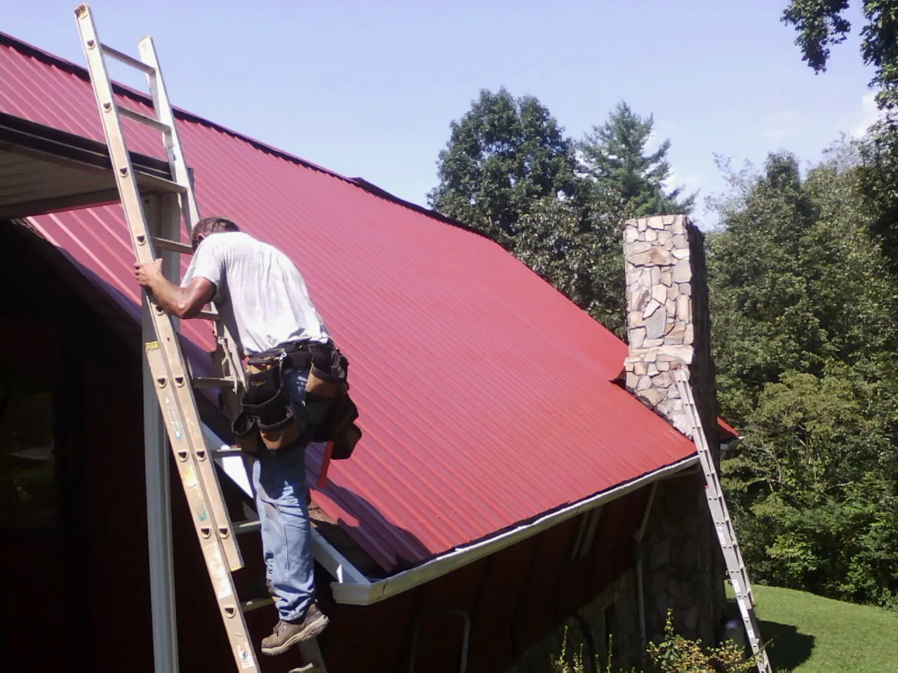 Roofer on a ladder beside a red metal roof and stone chimney — Soffit & Fascia Repair by Roofing Brothers in Alcalde